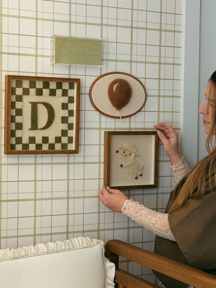Pamela pendurando o quadro do cordeirinho, ao lado do quadriculado com a letra D e do balão