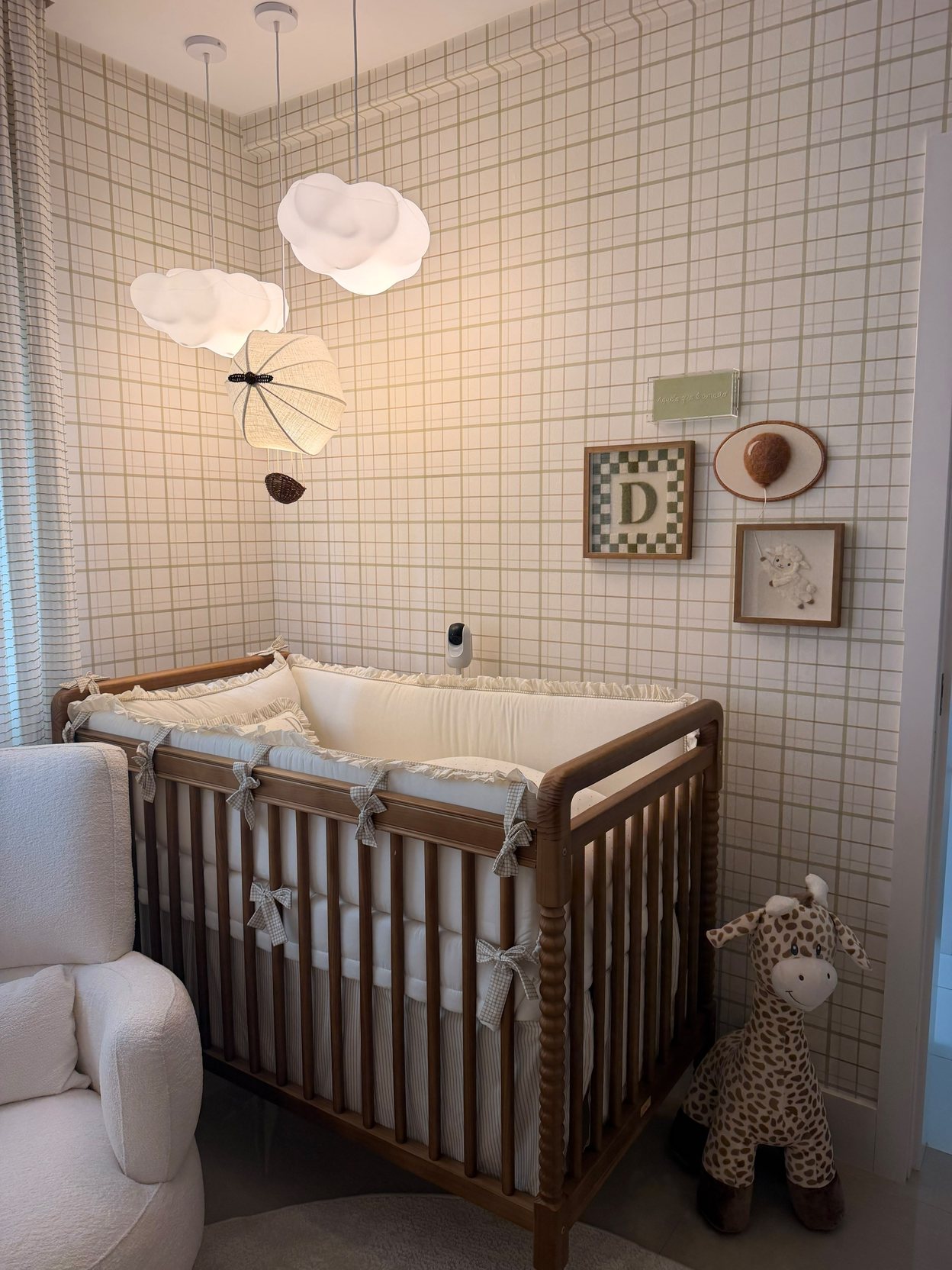 Quarto de bebê com berço de madeira; na parede quadriculada, o quadro da letra D, um balão e um cordeirinho bordados