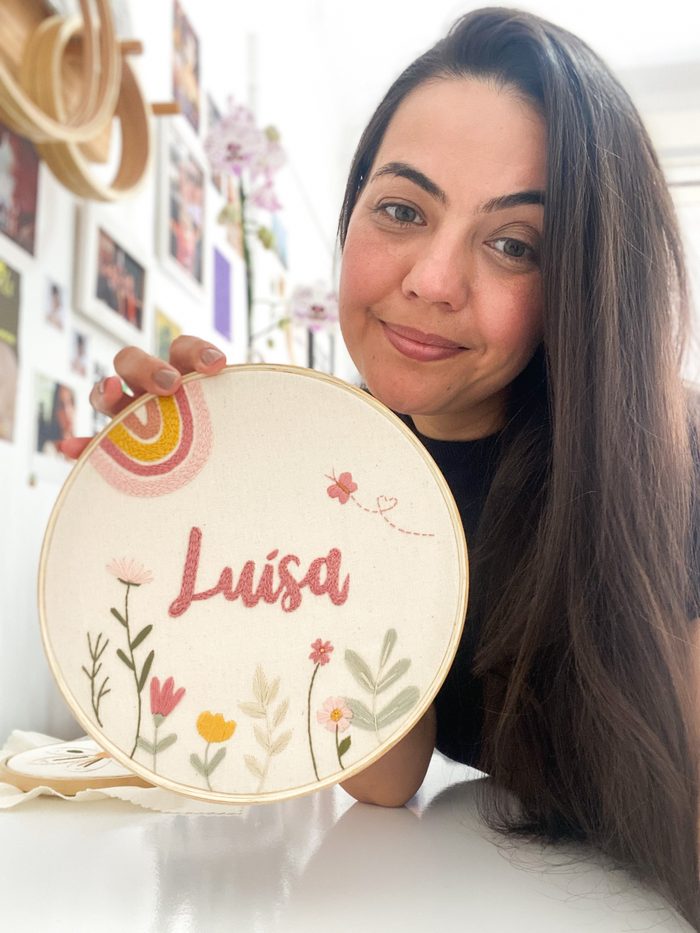 Pamela segurando o bastidor Luísa, com flores e arco-íris bordados